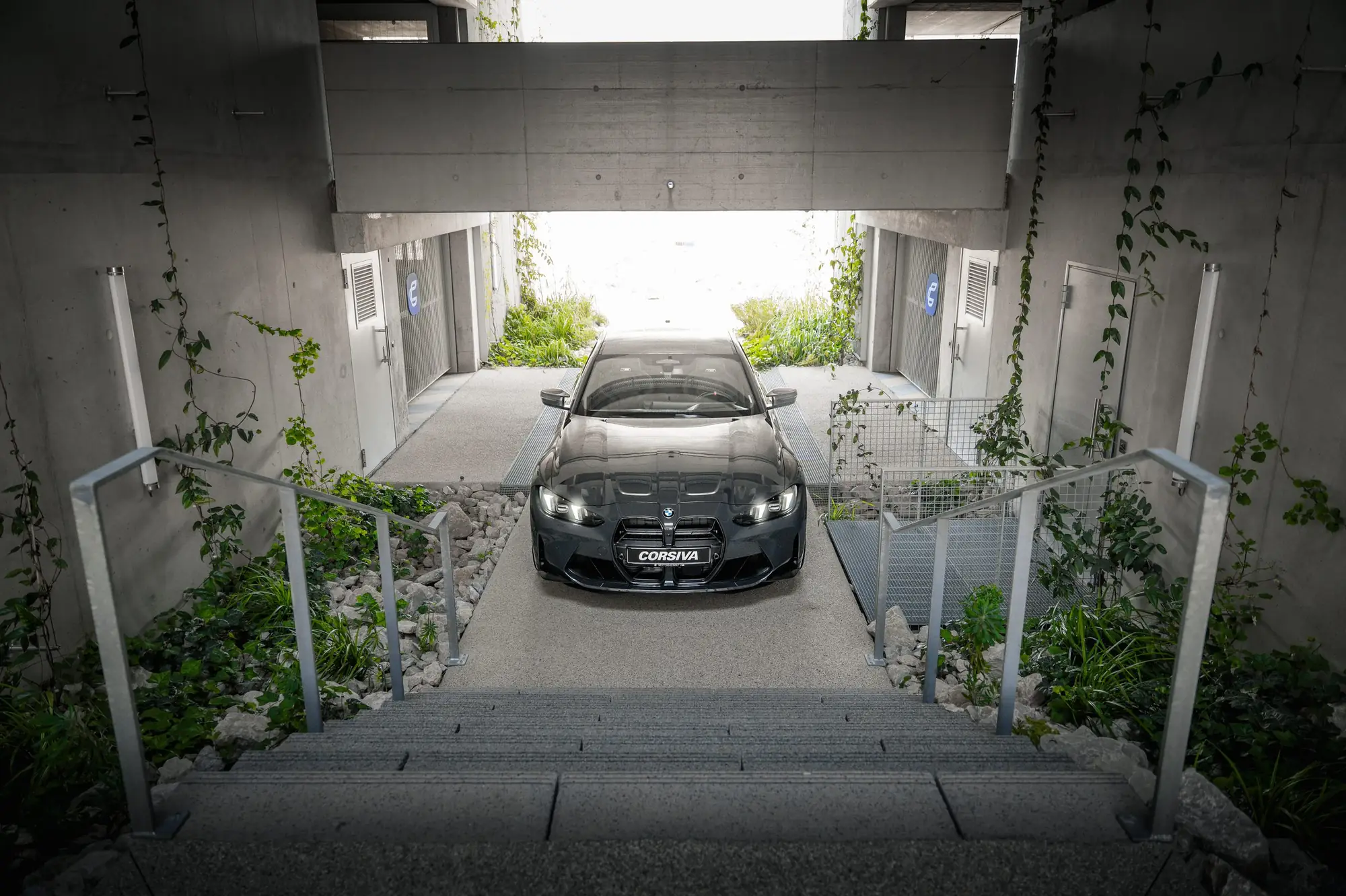 BMW M3 Competition du groupe Corsiva au pied des escaliers du garage, phares allumés