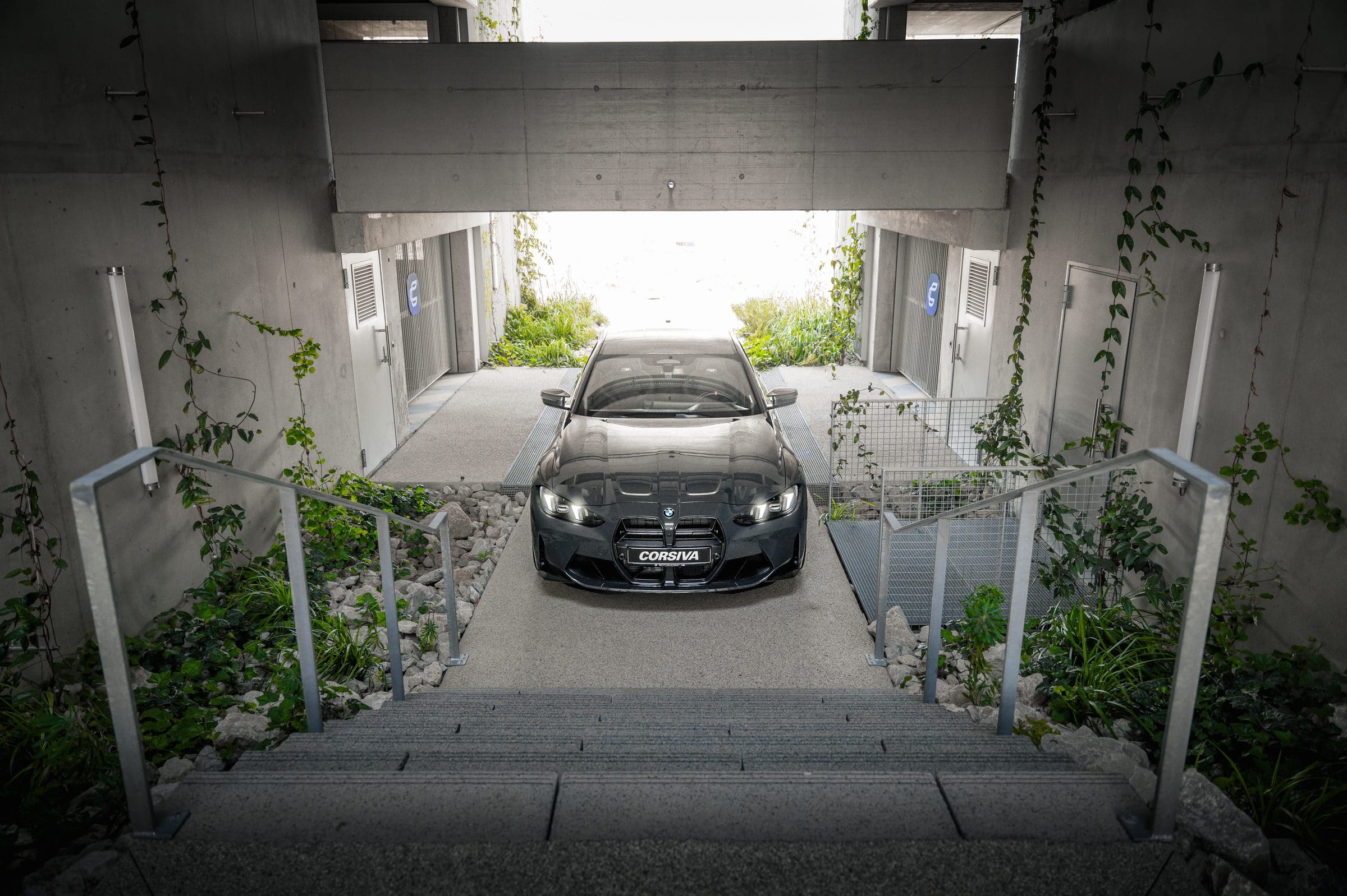 BMW M3 Competition du groupe Corsiva au pied des escaliers du garage, phares allumés