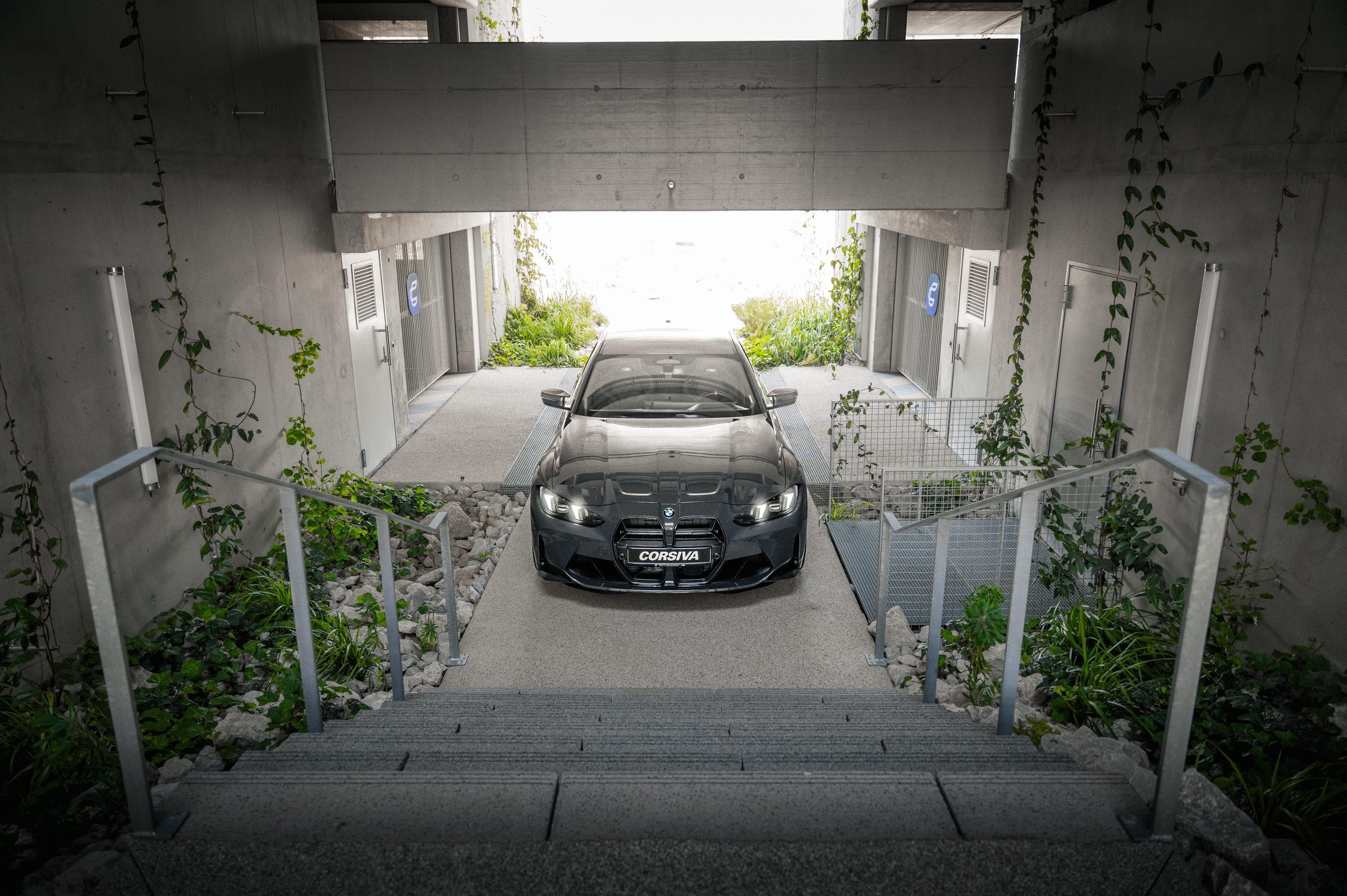 BMW M3 Competition du groupe Corsiva au pied des escaliers du garage, phares allumés